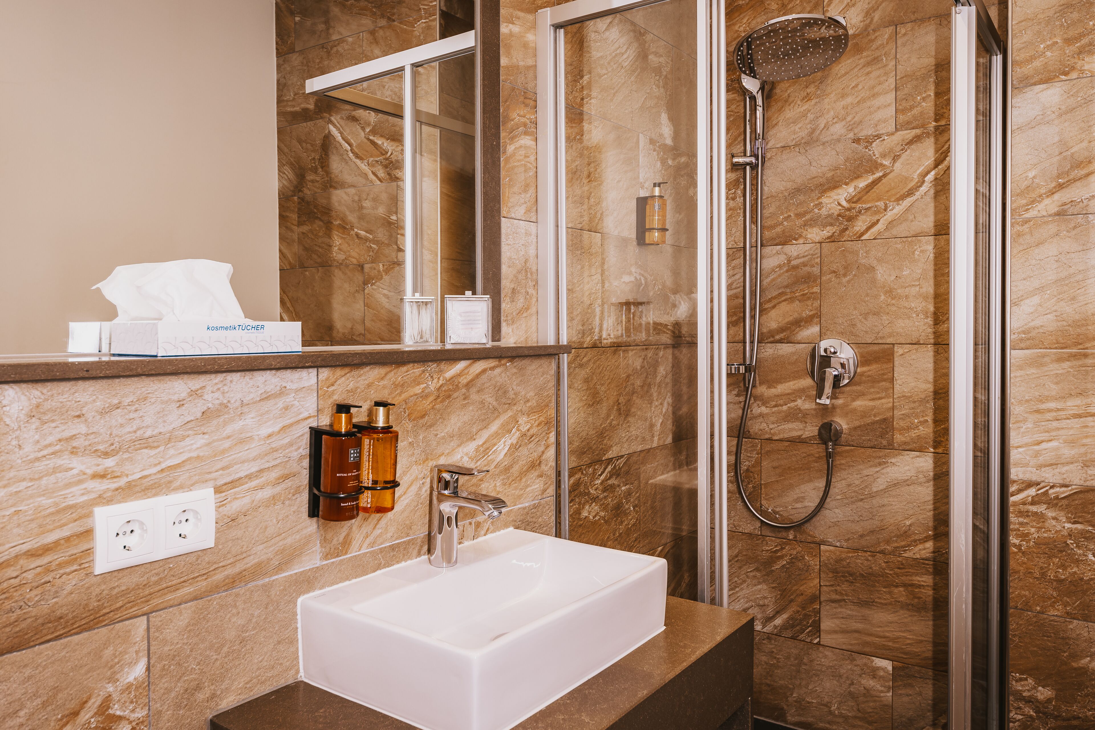 Un baño moderno con un lavamanos blanco, una cabina de ducha de cristal y paredes de mármol.