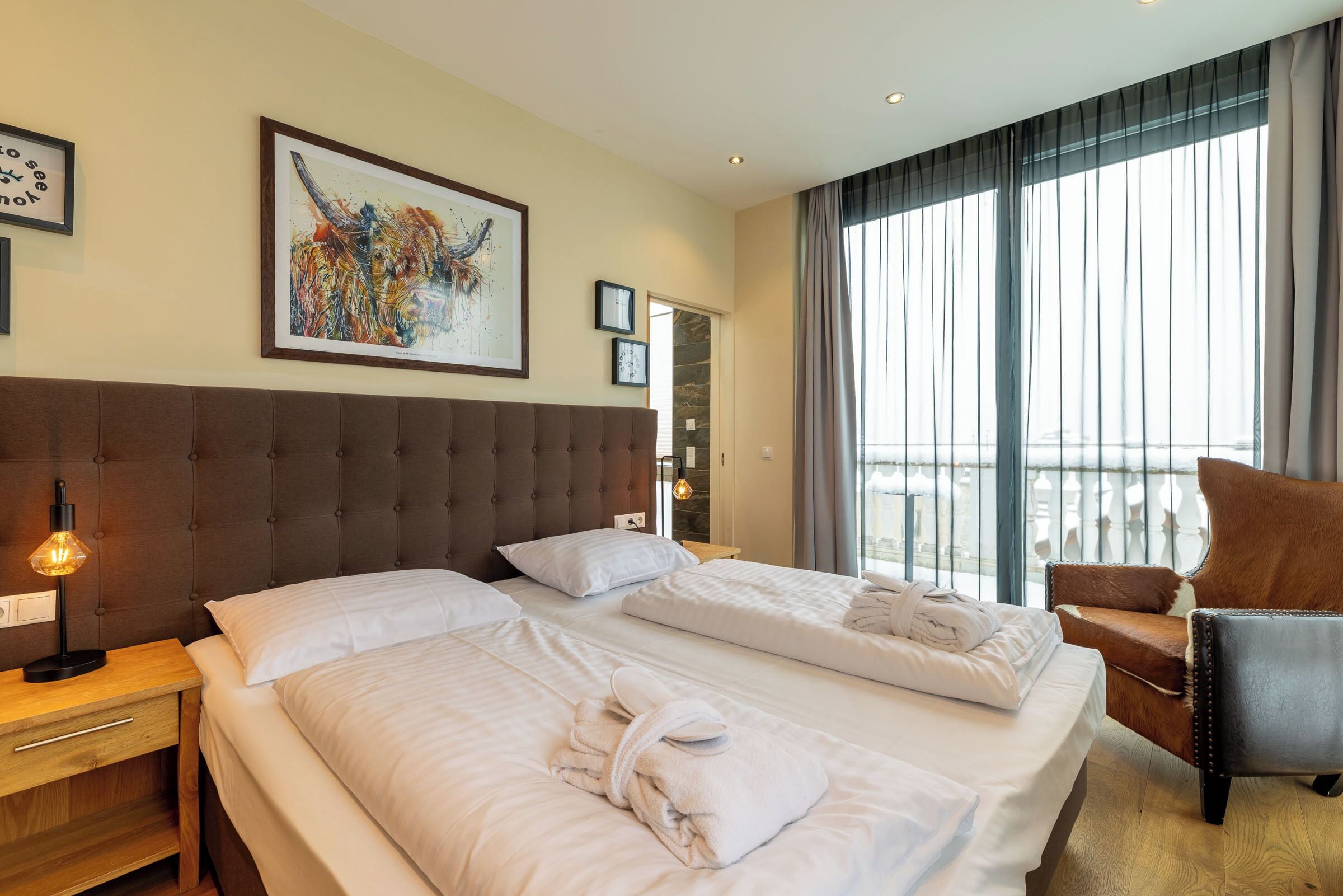 A hotel room with two beds, a large window with curtains, a brown upholstered headboard, and a brown leather chair.