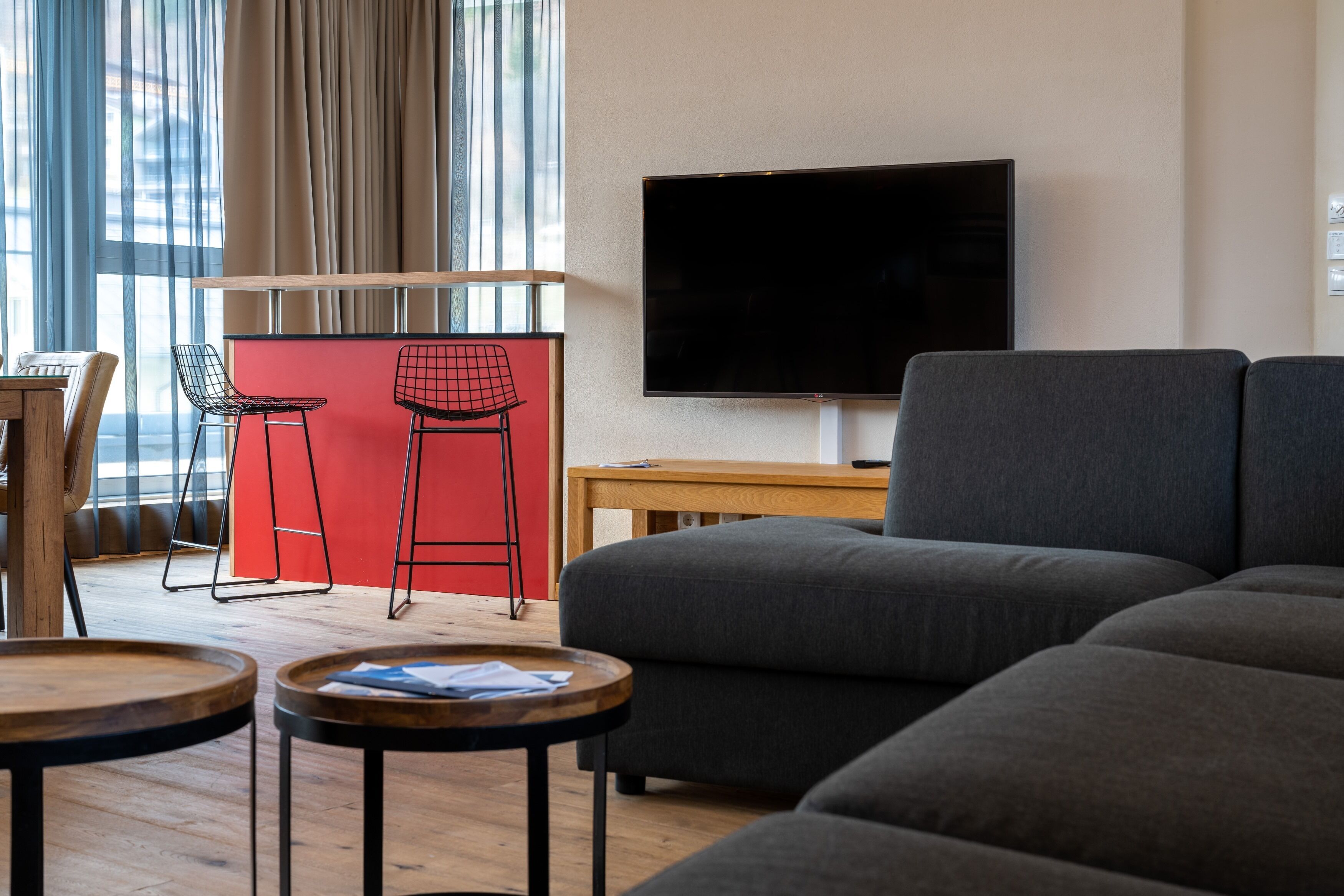 En moderne stue med et stort fladskærms-tv, en grå sofa, to røde barstole og et træspisebord.