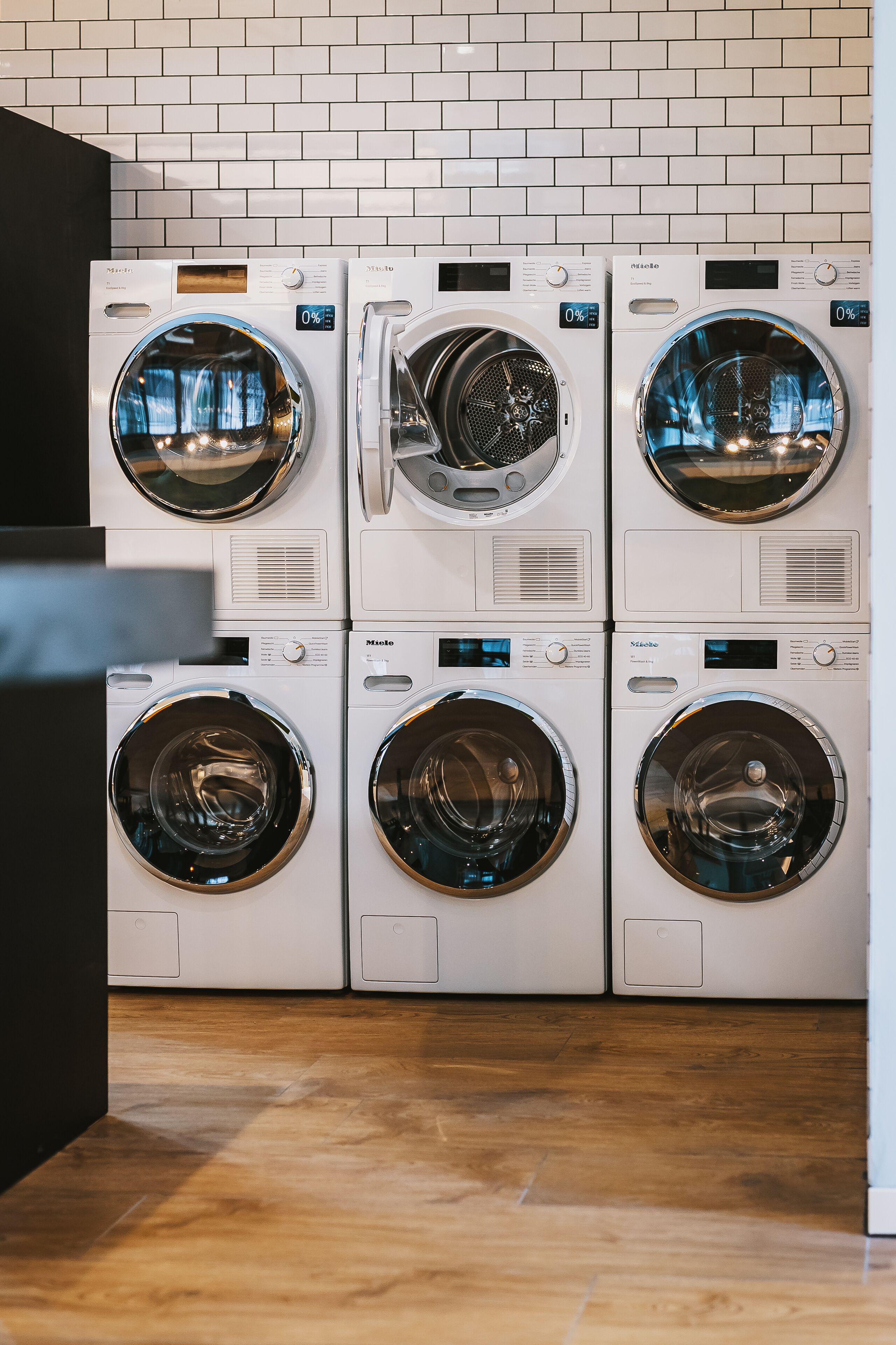 A row of six front-loading washing machines against a white tiled wall.