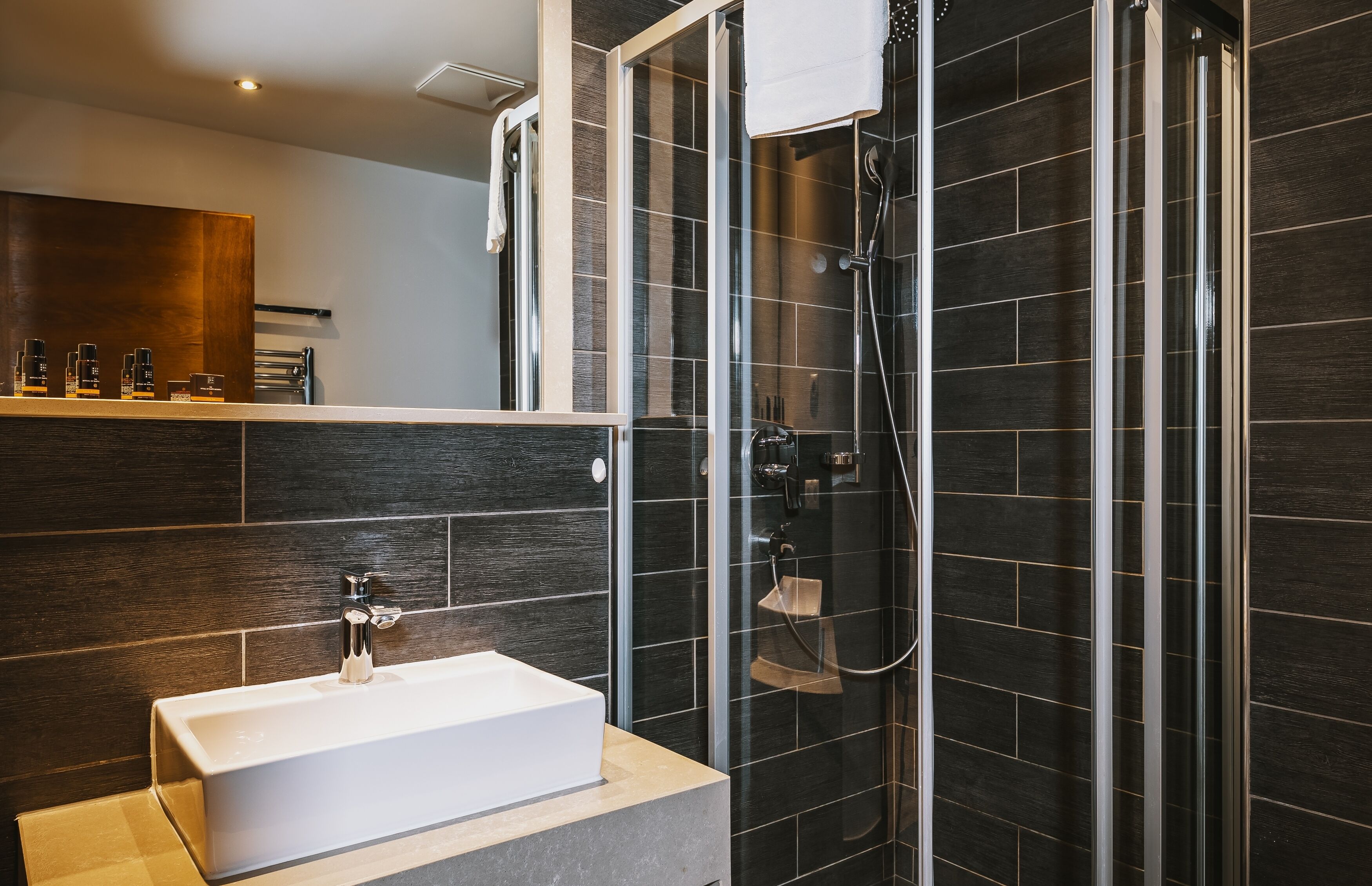 Un bagno moderno con cabina doccia in vetro, lavabo bianco e pareti piastrellate di colore scuro.