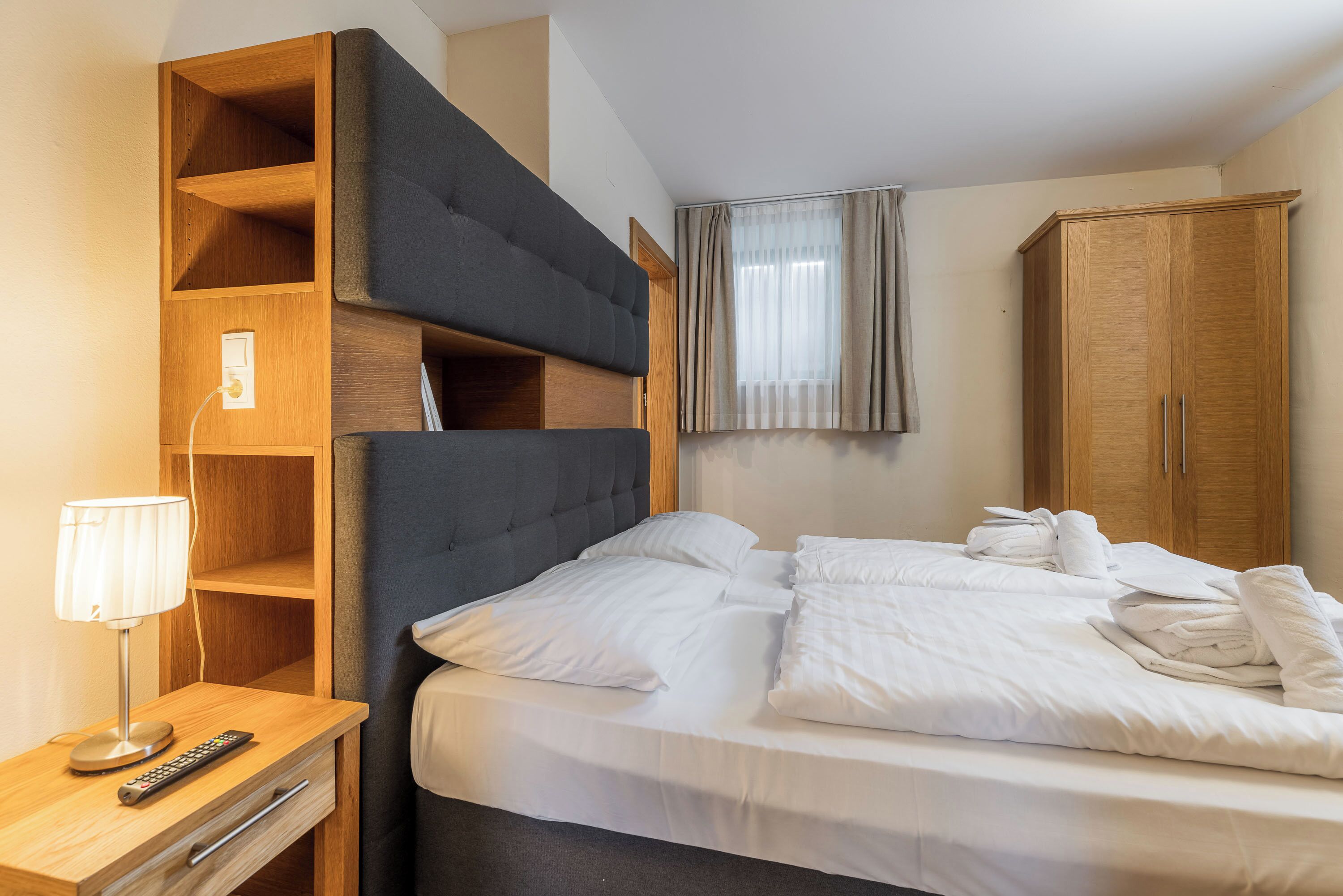 A hotel room with a bed, a nightstand with a lamp, a remote control, a wooden wardrobe, and a window with curtains.