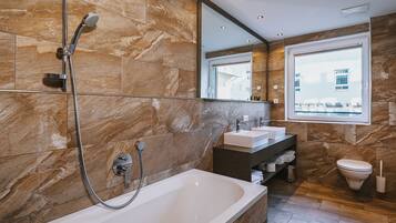 A modern bathroom with a large bathtub, two sinks, a toilet, and a window providing a view of buildings outside.