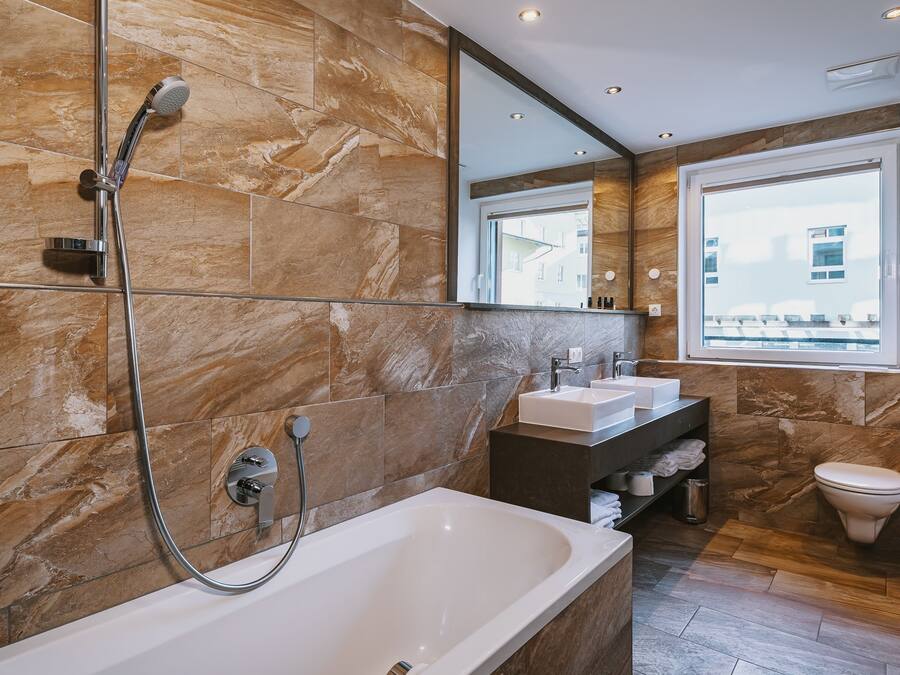 A modern bathroom with a large bathtub, two sinks, a toilet, and a window providing a view of buildings outside.