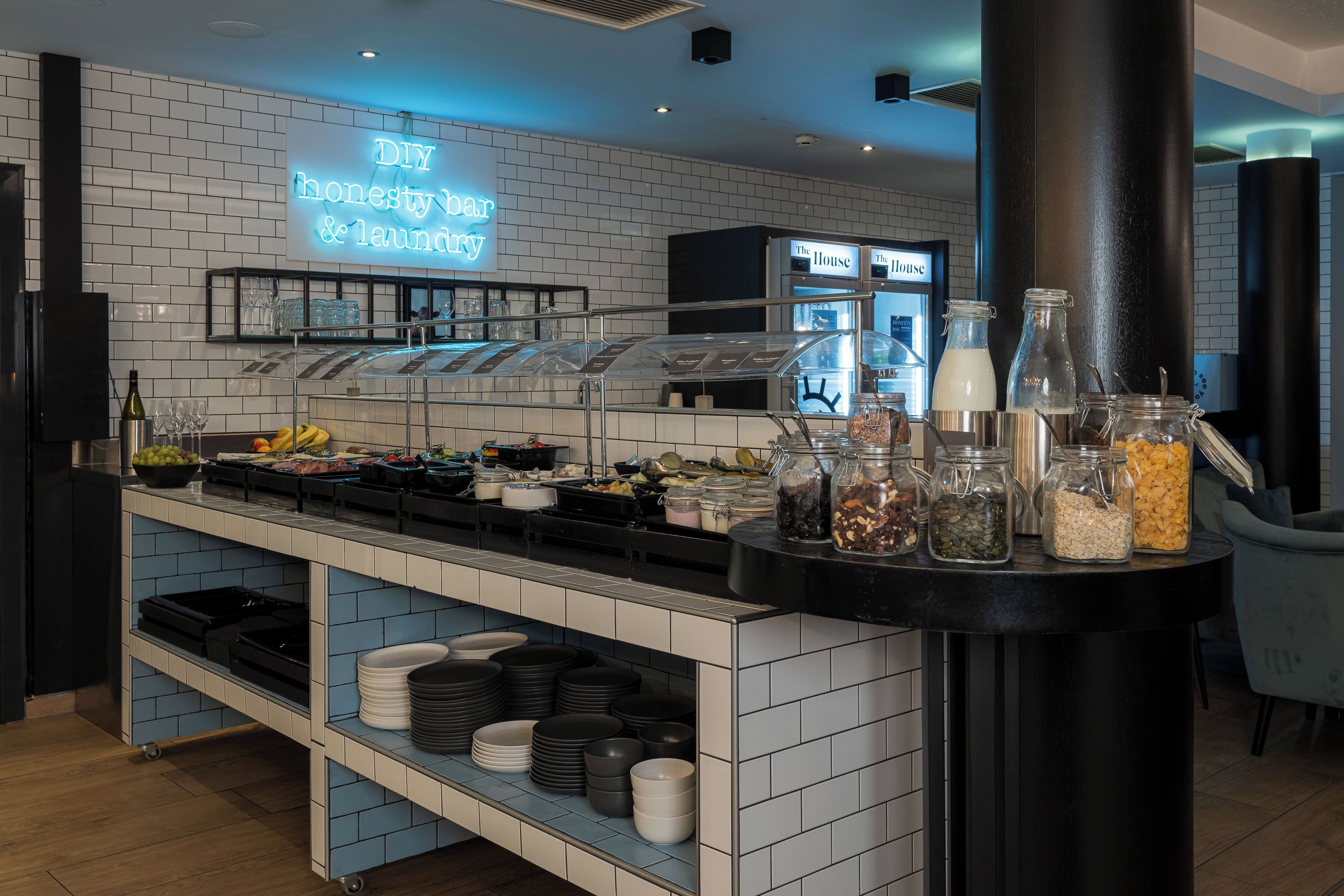 Ett område med en buffé, en neonskylt med texten ”Självbetjäningsbar med ärlighetsprincipen och tvättservice”, glasburkar med olika matvaror och ett urval av tallrikar och skålar på hyllor.
