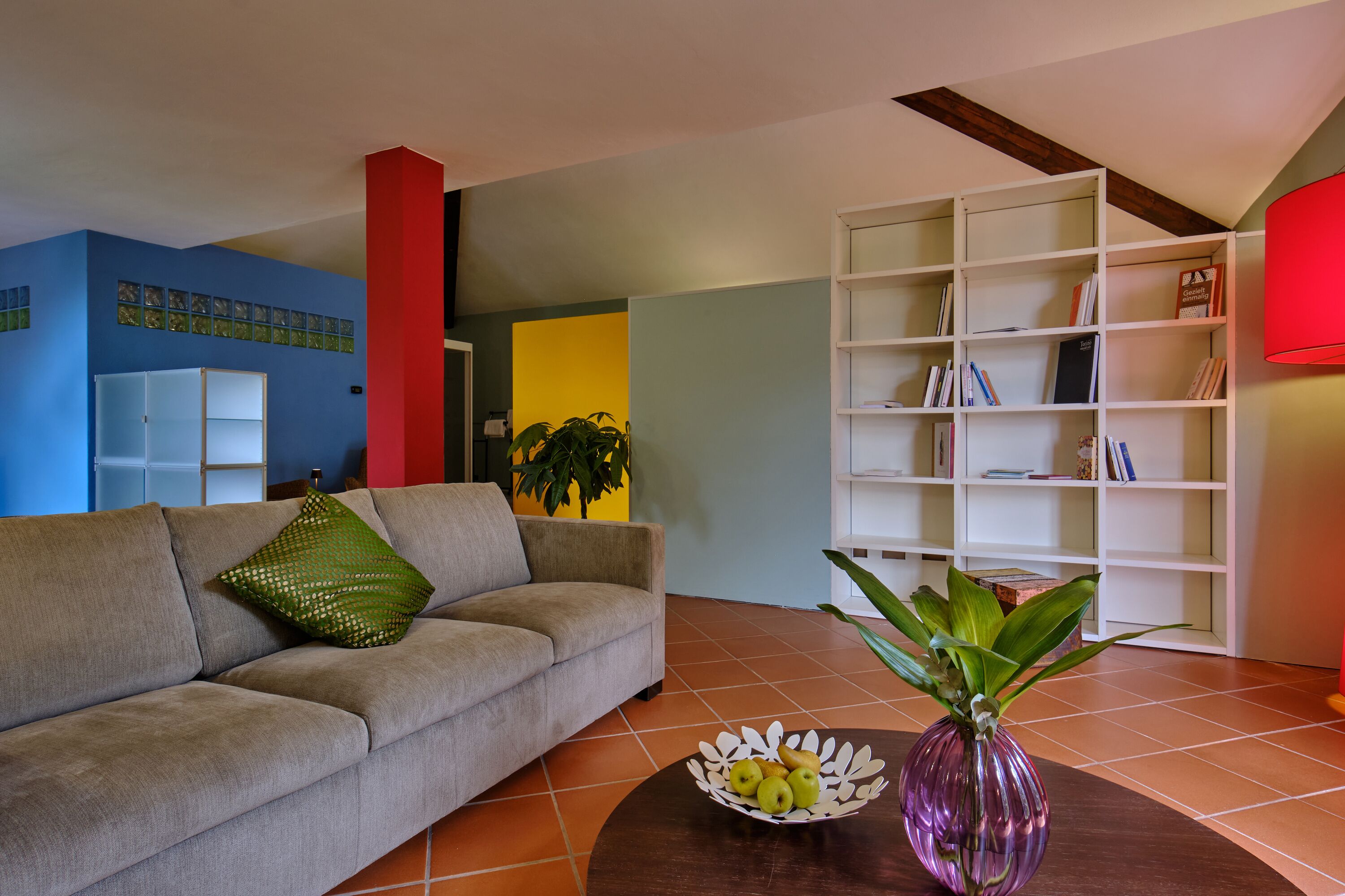 A living room with a sofa, a coffee table with a vase and fruit bowl, a bookshelf, and a red accent wall.