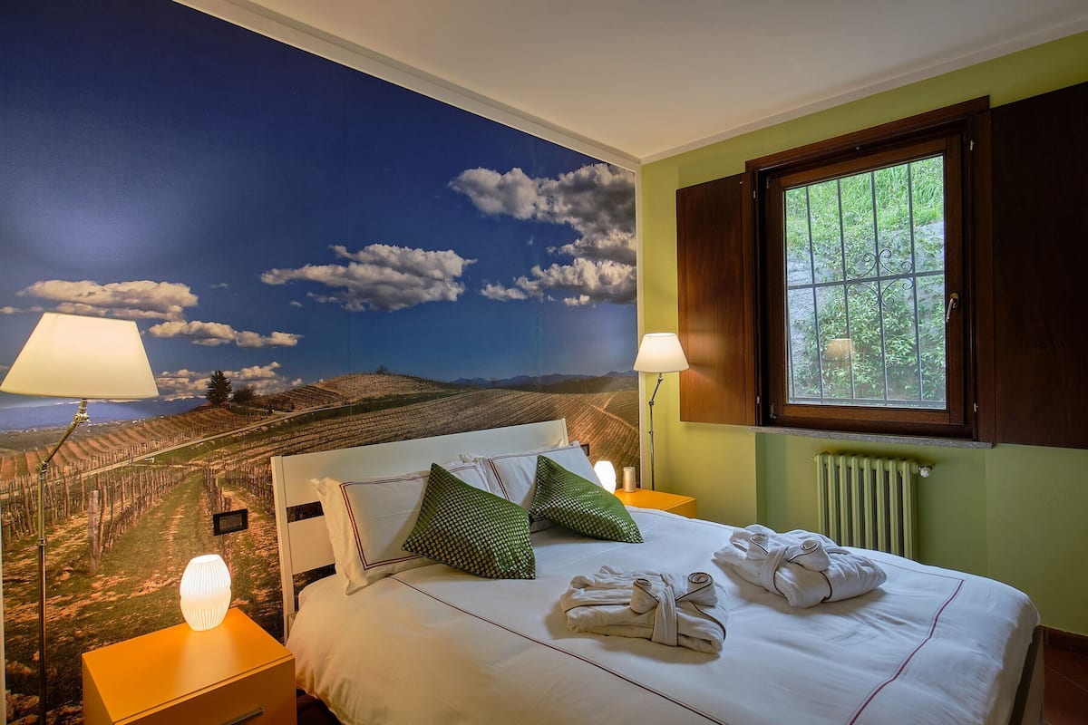 Hotelkamer met een muurschildering van een wijngaard, een netjes opgemaakt bed met kussens, twee bedlampen en een raam met gordijnen.