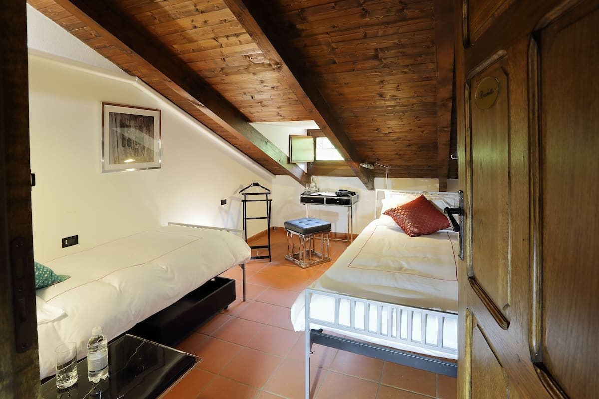 Een slaapkamer met twee bedden, een houten plafond, een klein krukje en een nachtkastje met een glas en fles.