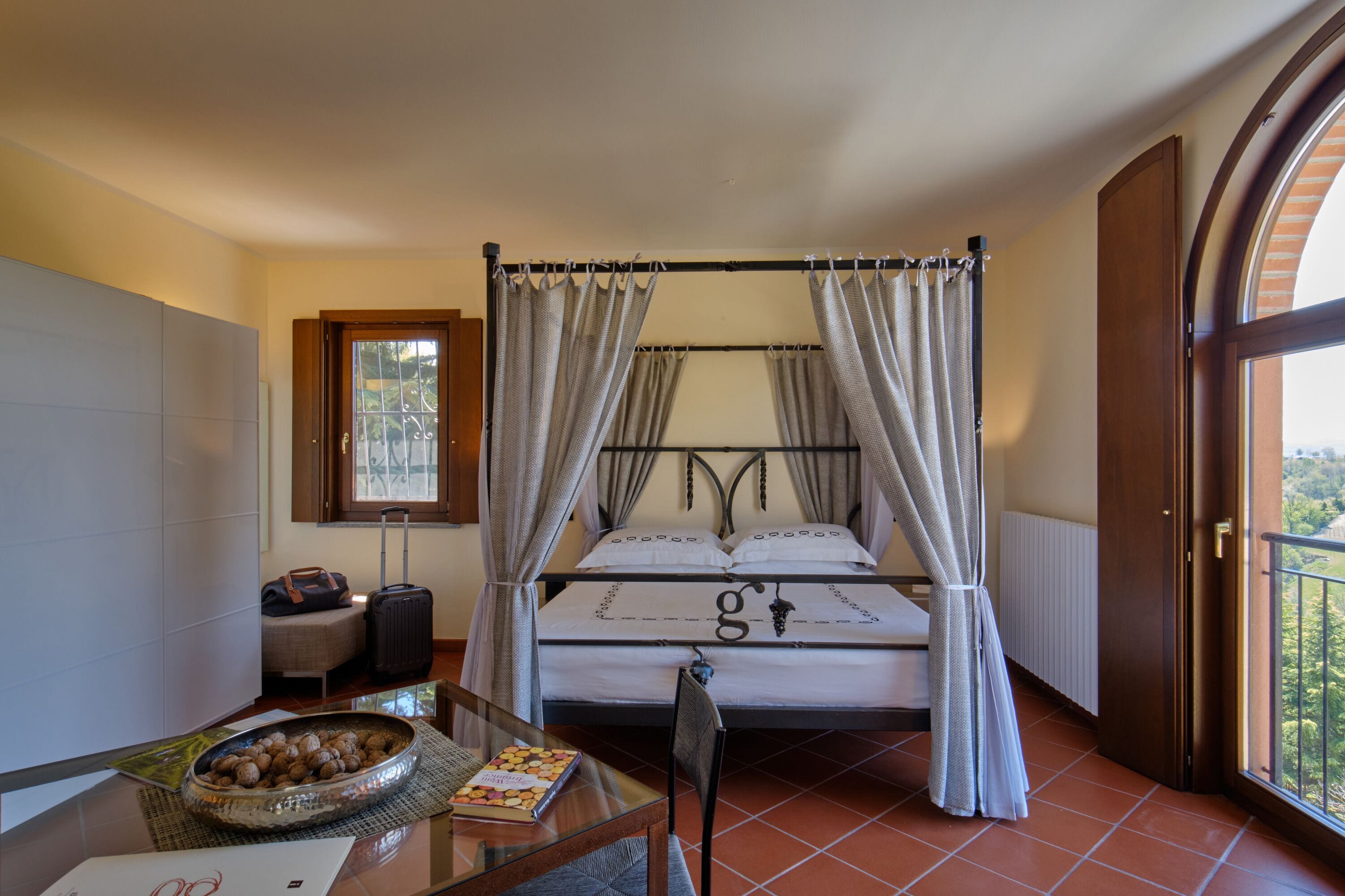 A four-poster bed with a canopy, a small table with a bowl of nuts, a chair, and a suitcase.