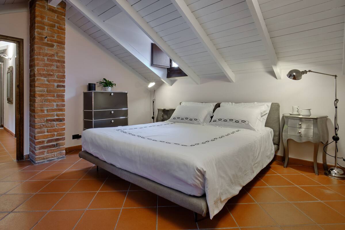 Een slaapkamer met een groot bed, een bakstenen muur, een plafond met zichtbare balken, en een klein nachtkastje met een lamp.