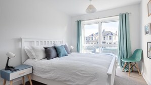 Une chambre à coucher avec un lit, une table de chevet, une lampe, une chaise, une fenêtre et des rideaux.