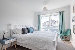 A bedroom with a bed, bedside table, lamp, chair, window, and curtains. - Spacious Flat, Sea Views & Private Parking, Fistral (Newquay)