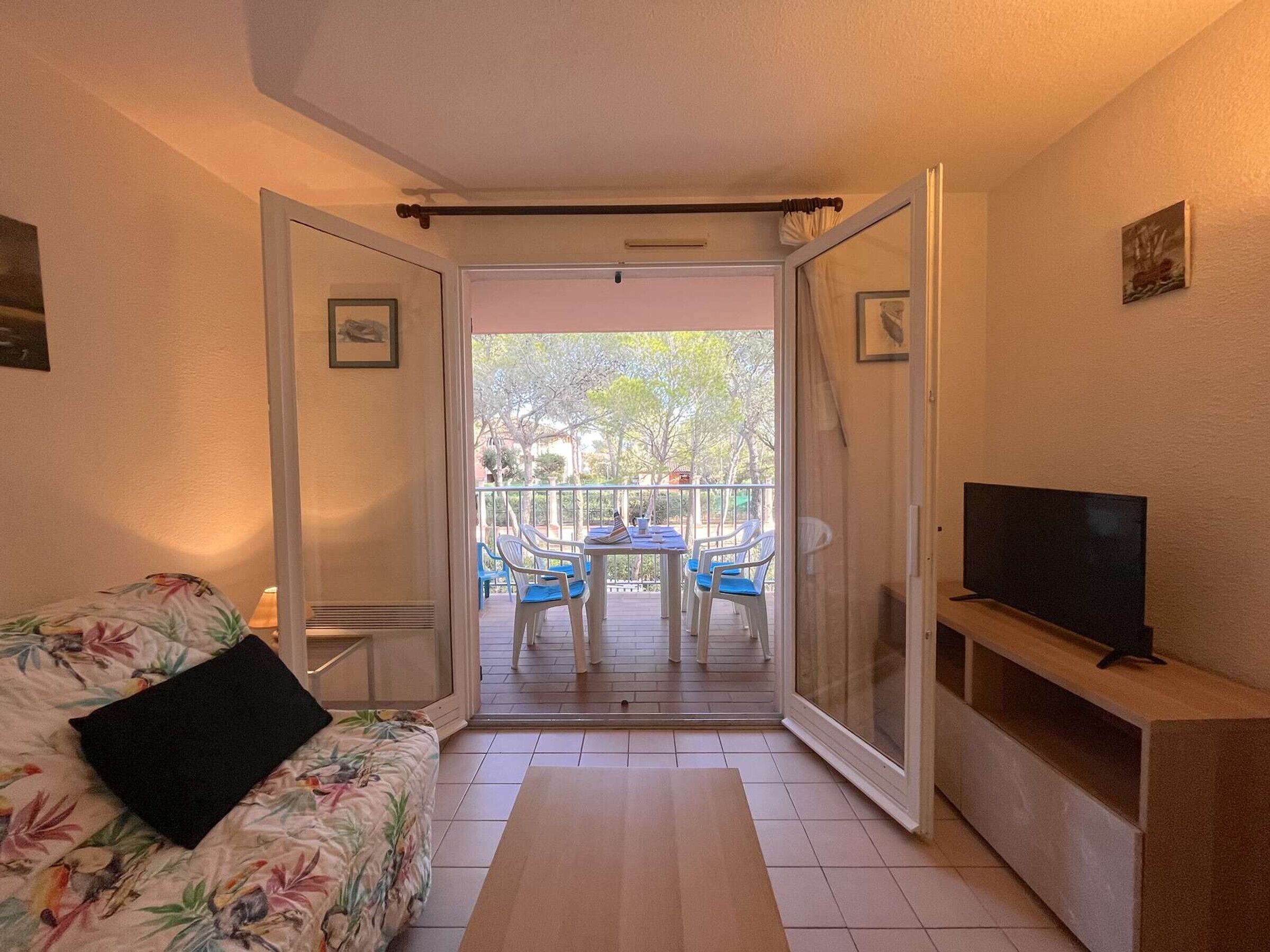 A room with a bed, a TV on a stand, a sofa, and a balcony with chairs and a table.