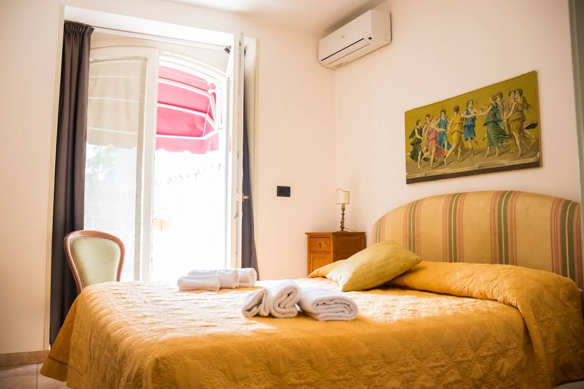 A hotel room with a bed, a chair, a painting, and an air conditioning unit.