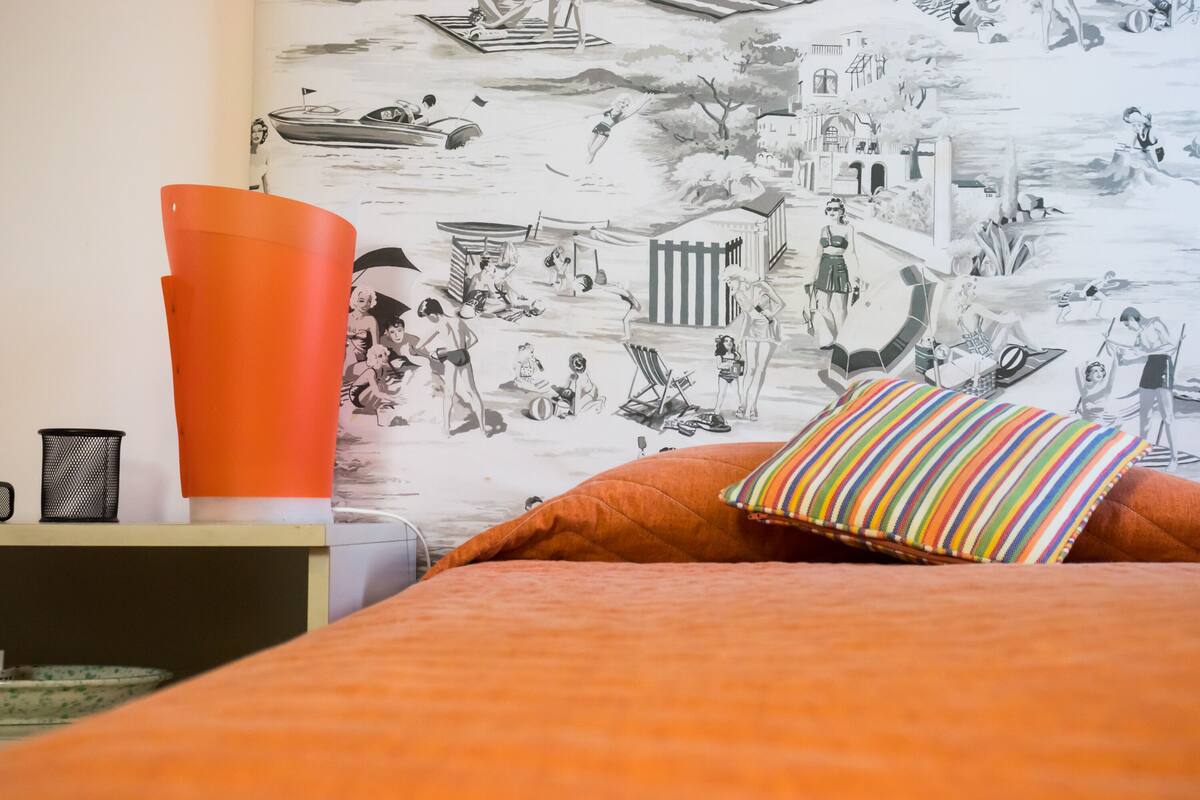 A bedroom with a mural of a beach scene, a bed with orange bedding, and a striped pillow.