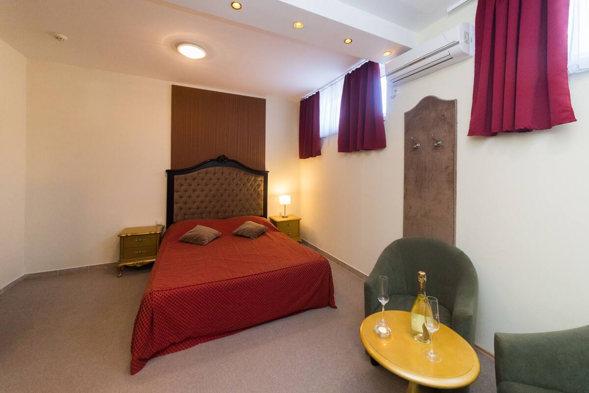 A hotel room with a bed, a nightstand, a lamp, a chair, and a table with a bottle and glass.