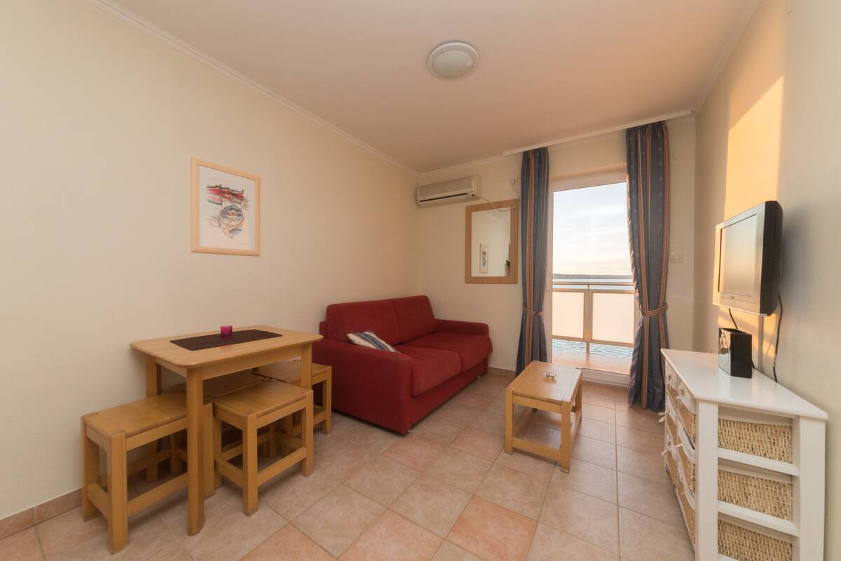A compact living room with a red sofa, wooden furniture, a TV, and a balcony with curtains.