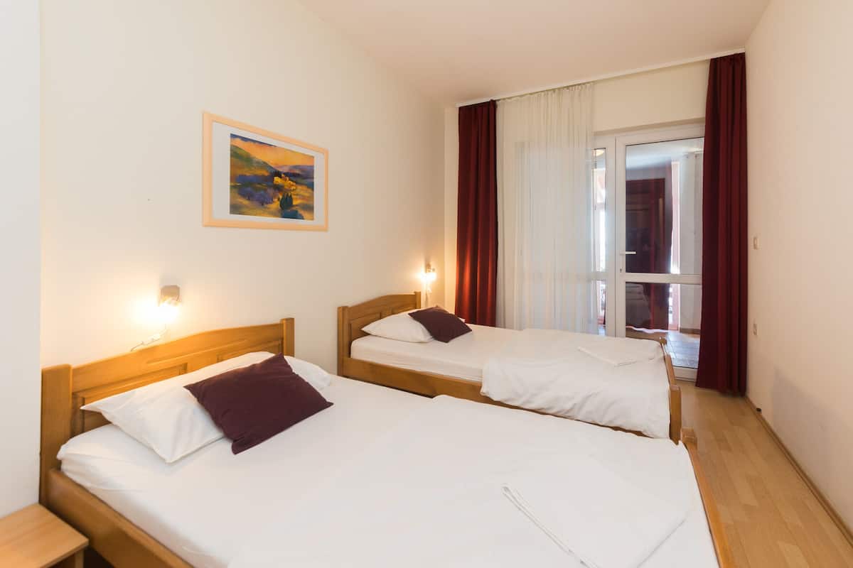 A hotel room with two single beds, a painting on the wall, and a view of the outside through a window with curtains.