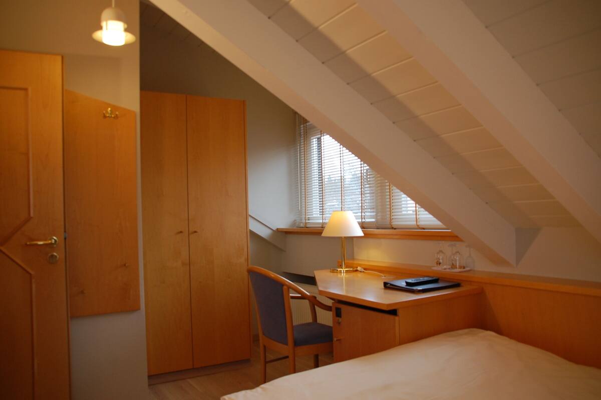 Een kamer met een bed, een bureau en een stoel. Er is een raam met zonwering en een lamp op het bureau.