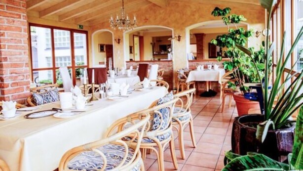 A dining area with wooden tables and chairs, potted plants, and a brick wall. - Sporthotel  Schönblick (Meersburg)