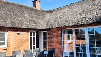A brick building with a thatched roof, a patio with outdoor seating, and a glass door leading to an interior space.