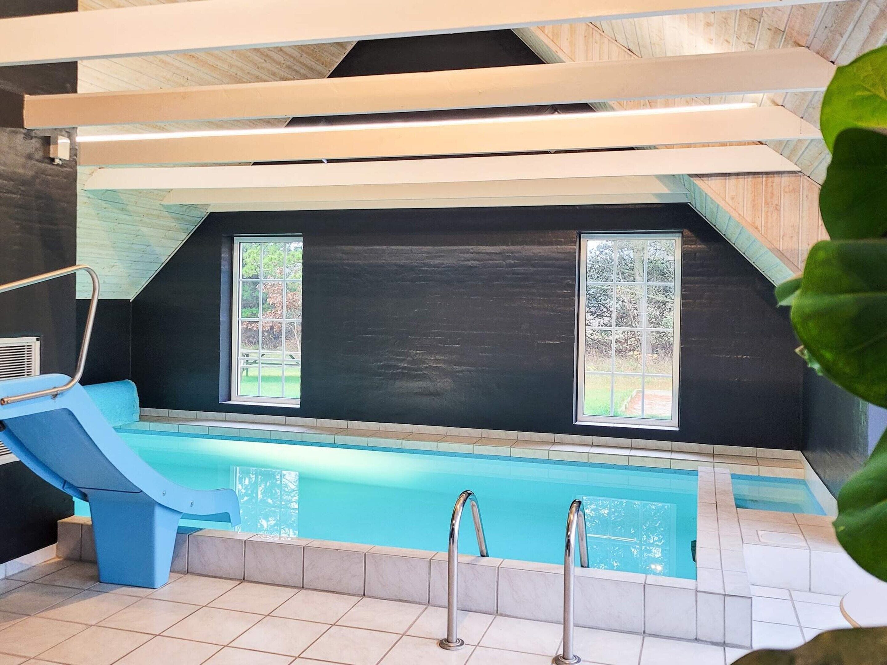 En indendørs swimmingpool med blå rutsjebane, to vinduer og fliset gulv.
