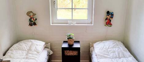 A room with two single beds, a small table with a plant, and a window with blinds.