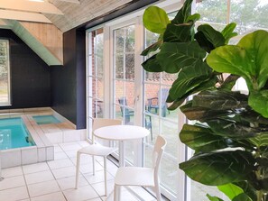 Uma área interna de piscina moderna com parede de vidro, duas cadeiras brancas e uma grande planta verde.