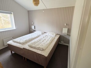 A single-bed room with a wooden headboard, a bedside table with a lamp, and a window with blinds.