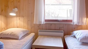 A room with wooden walls, two beds, a small table, a window with white curtains, and a wall-mounted light fixture. - Seaside Escape in Snogebaek - By Traum Ferienwohnungen (Nexø)