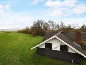 A small, single-story house with a sloped roof, a front porch, and a view of a grassy field and trees.