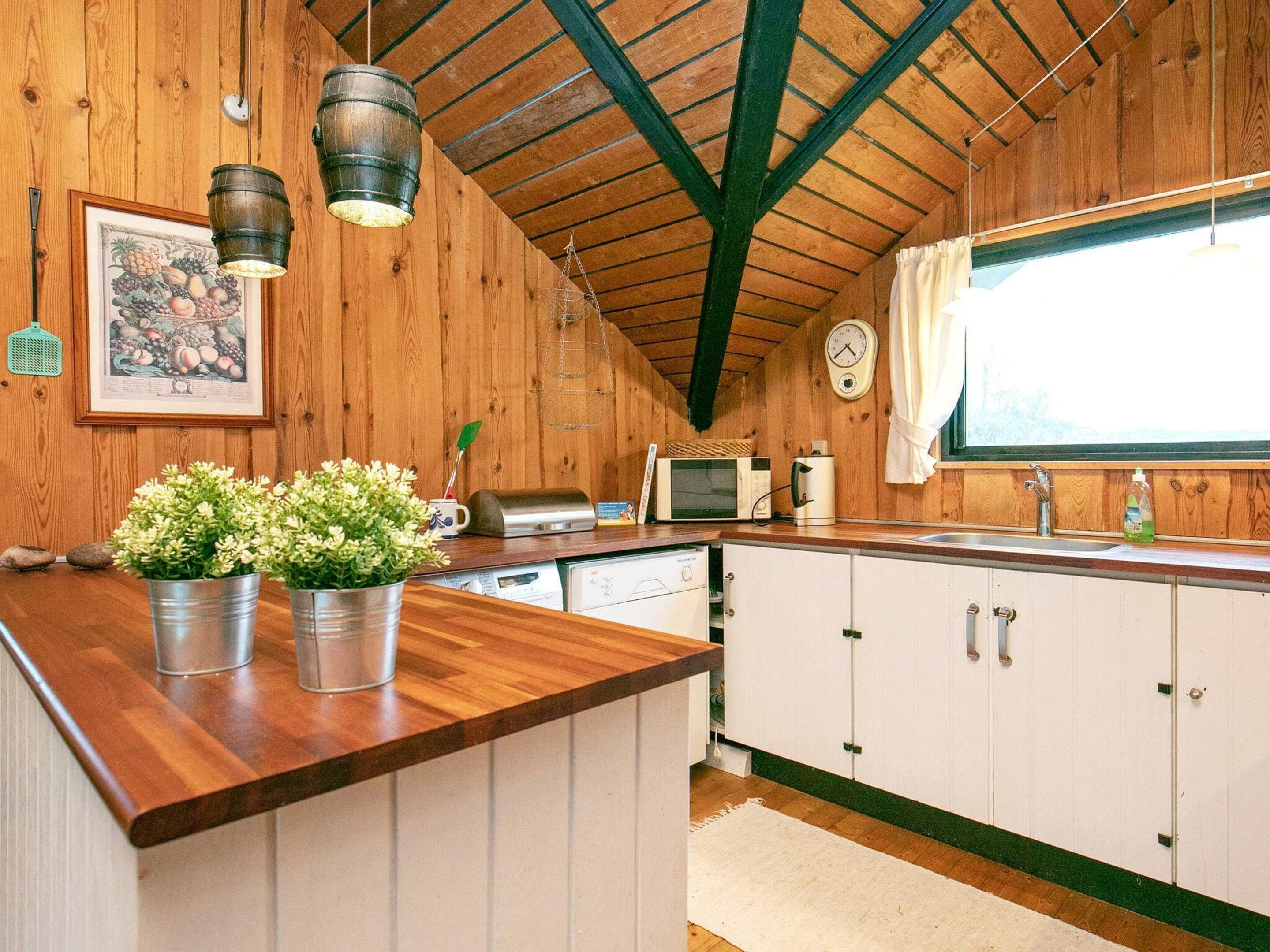 A kitchen with wooden walls, a wooden ceiling with green beams, a wooden countertop, white cabinets, a microwave, a toaster, a sink, a clock, and a window with curtains.