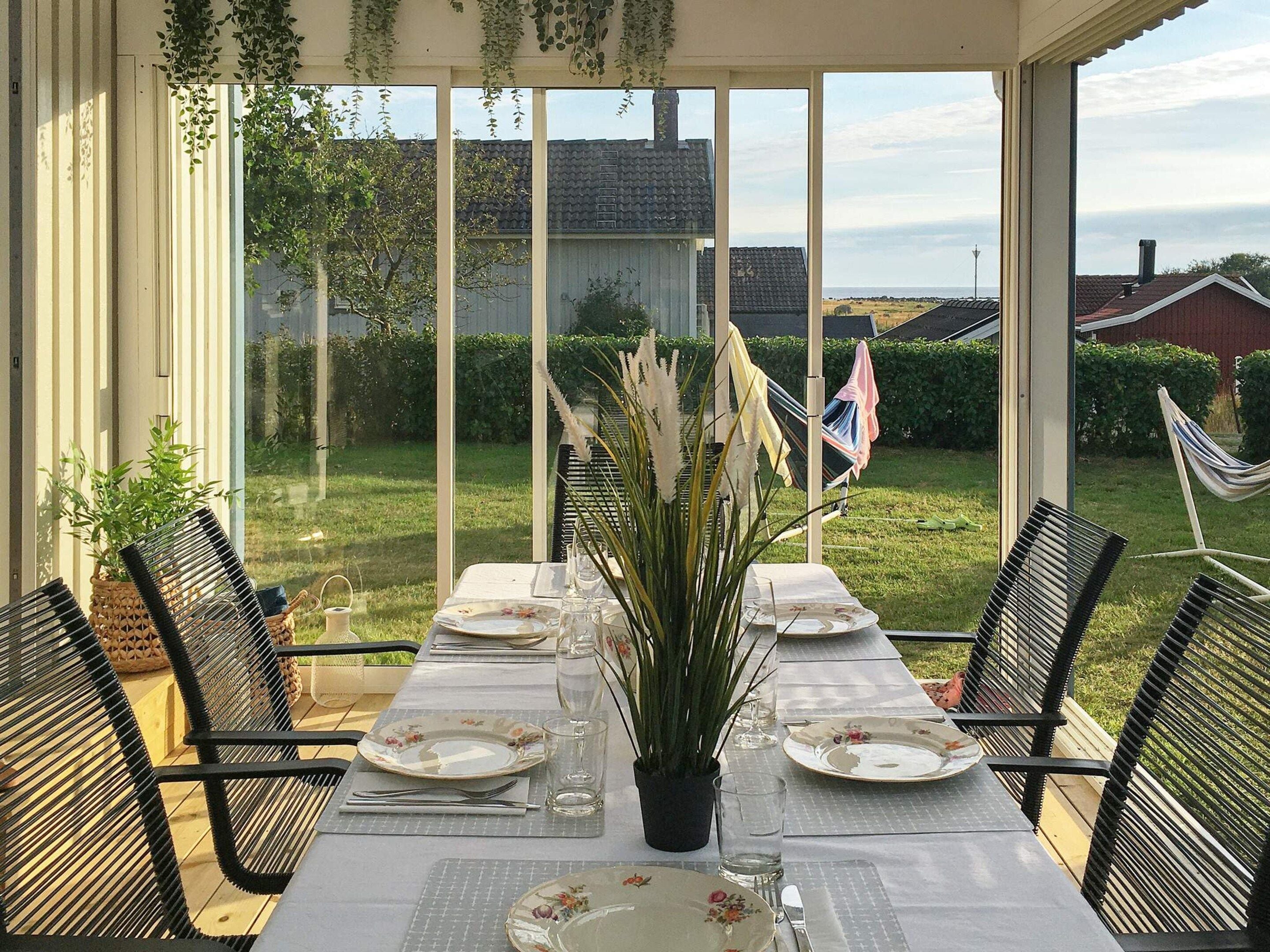An outdoor dining area with a table set for four, surrounded by chairs, with a view of a house and garden in the background.