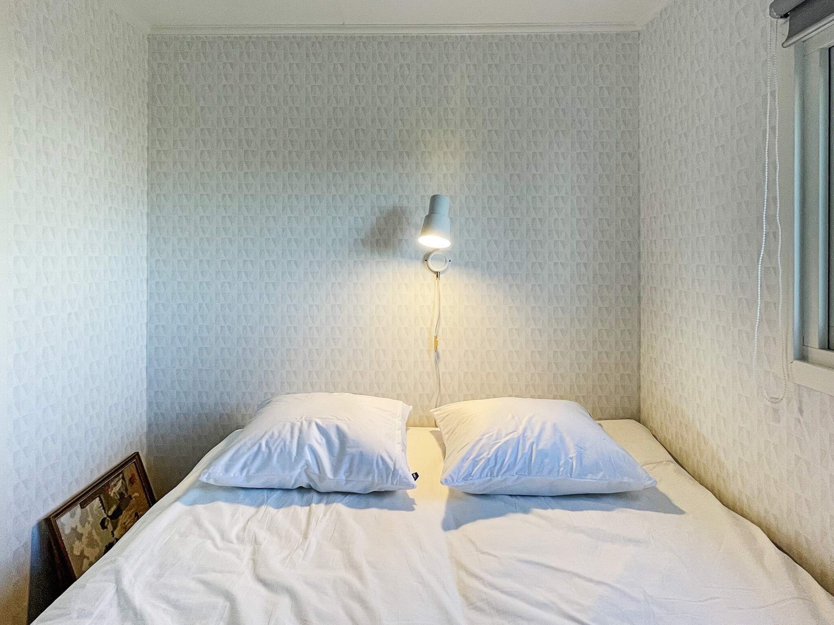 A single bed with white bedding and two pillows, a wall-mounted light fixture, and a framed picture on the left side.