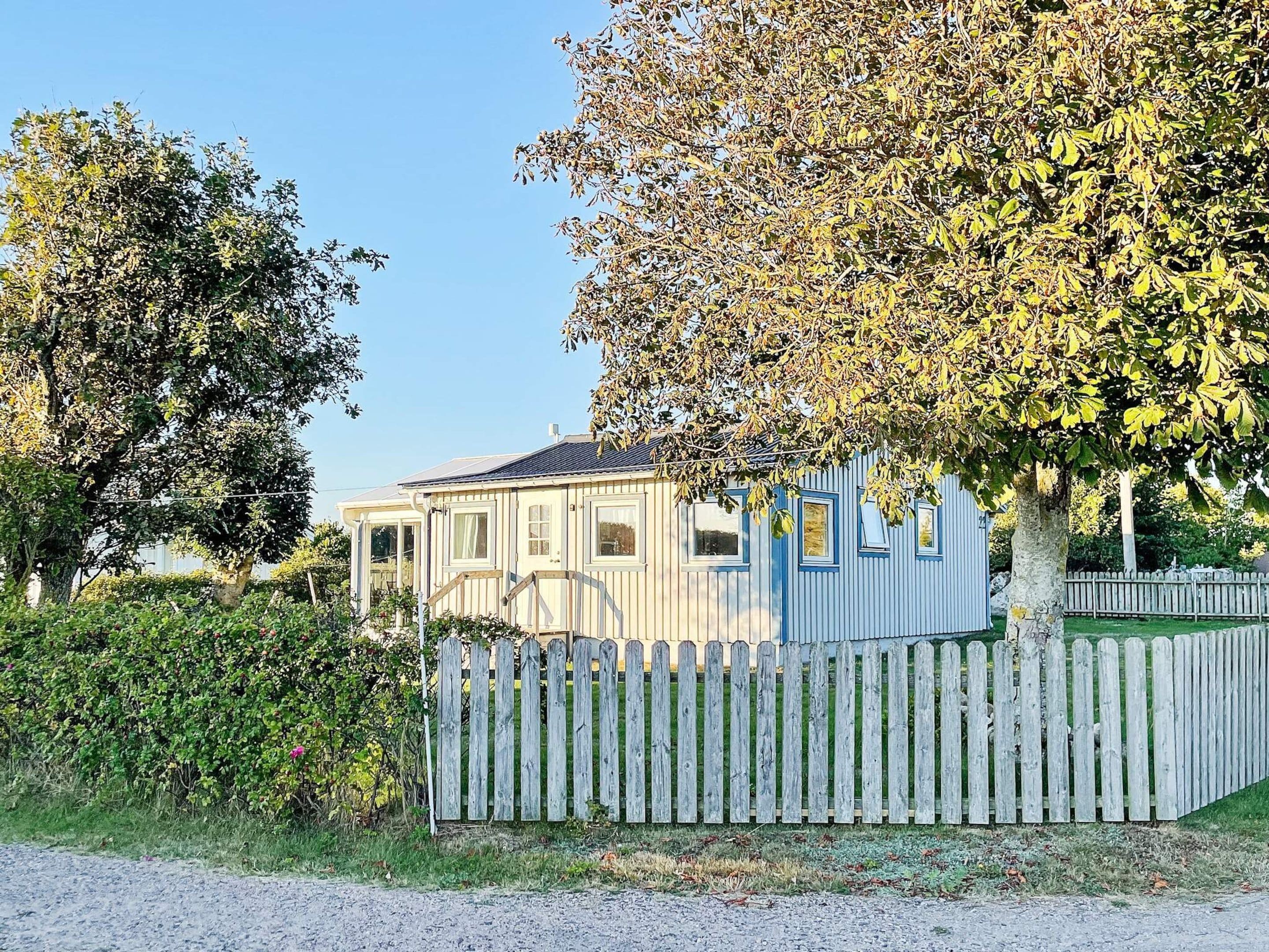 A single-story house with a wooden fence, surrounded by trees and a clear sky.