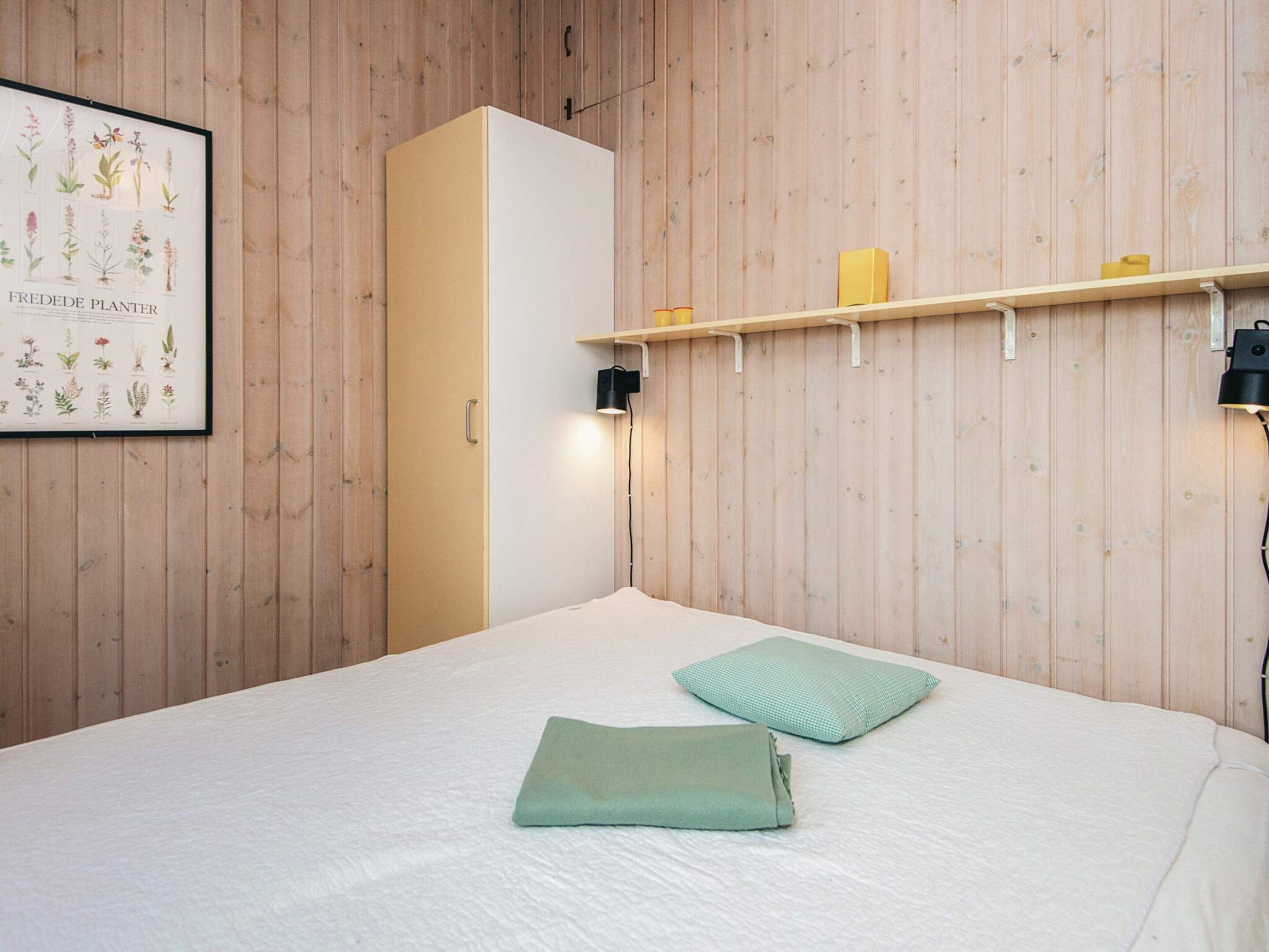 A bedroom with a wooden wall, a bed with a white sheet, a green pillow, a framed botanical print, and a shelf with candles and a lamp.