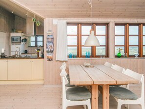A modern kitchen with wooden cabinets, a dining table with white chairs, and large windows with a view of the outdoors. - 4 Star Holiday Home in Hvide Sande (Hvide Sande)
