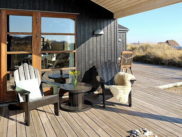 A wooden deck with Adirondack chairs, a round table, and a glass door leading to a room.