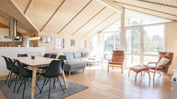 A spacious loft with high ceilings, a dining area, a living room with a sofa and armchairs, and large windows offering a view of the outdoors.