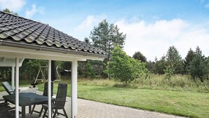 A covered outdoor seating area with a table and chairs, a swing set, and a grassy lawn with trees in the background.