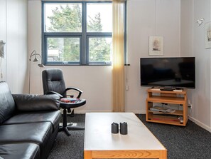 A living room with a leather sofa, a black office chair, a television on a wooden stand, and a window with a view of trees. - 6 Person Holiday Home in Hvide Sande (Hvide Sande)