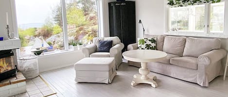 A living room with a fireplace, a sofa, a coffee table, and large windows with a view of greenery.