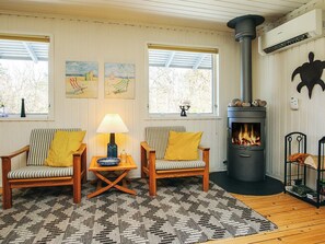 Una sala de estar acogedora con dos sillones a rayas, una mesa de centro de madera y un tapete con estampado blanco y negro. Se ve una chimenea con una chimada negra, y hay dos ventanas con obras de arte encima.
