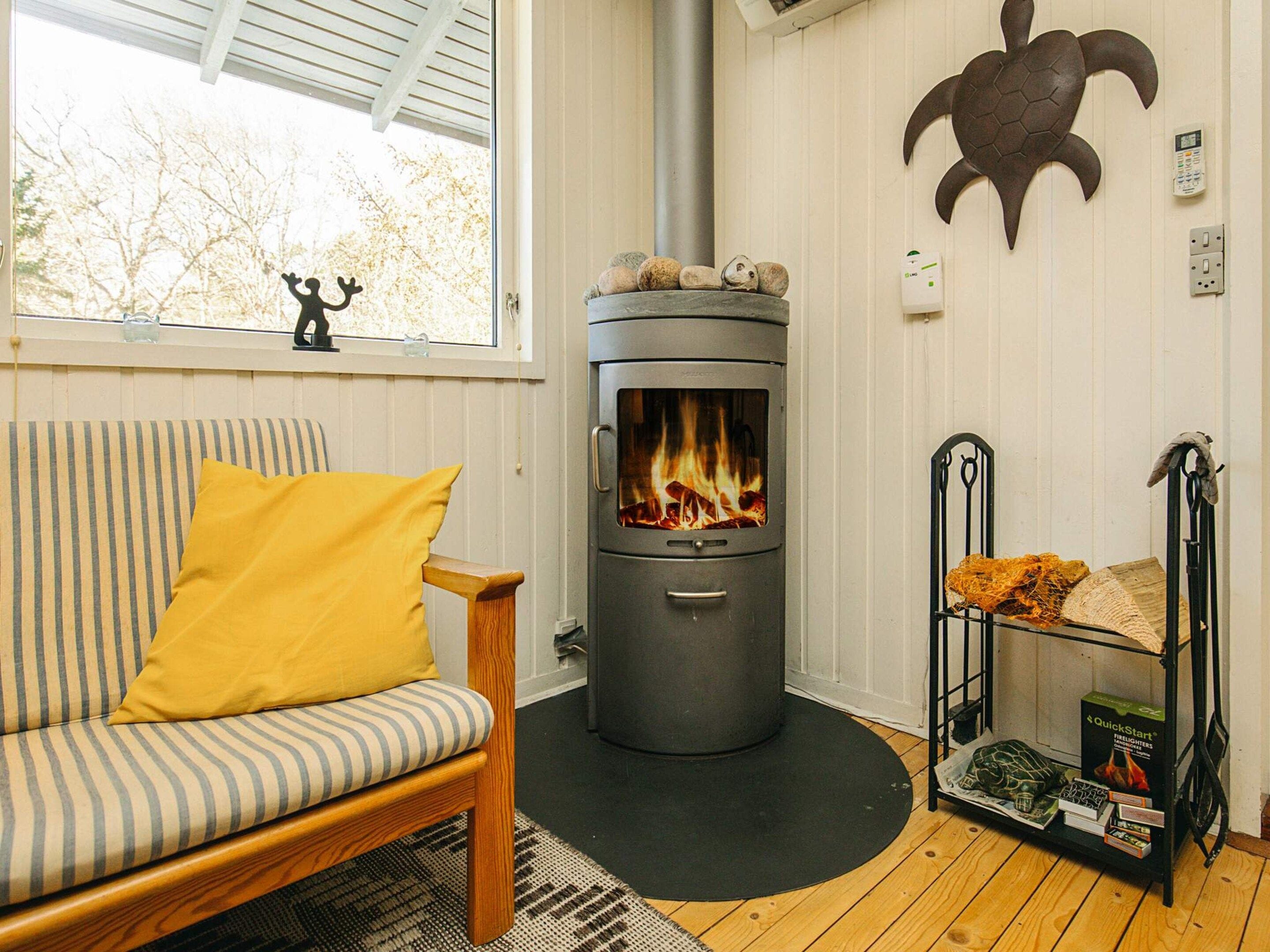 A cozy room with a wood-burning stove, a striped sofa with a yellow pillow, and a shelf with firelighters.