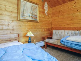 A wooden cabin room with a bed, a chair, a table with a lamp, and a painting on the wall.
