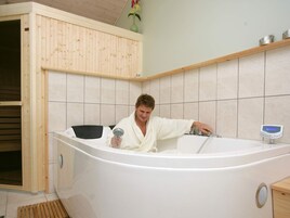 A person in a white bathrobe sitting in a large bathtub with a digital control panel, wooden cabinetry, and a tiled wall.