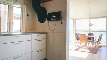 Et moderne køkken med hvide skabe, en hængende sort genstand og udsigt til et udendørs spiseområde med et bord og stole.
