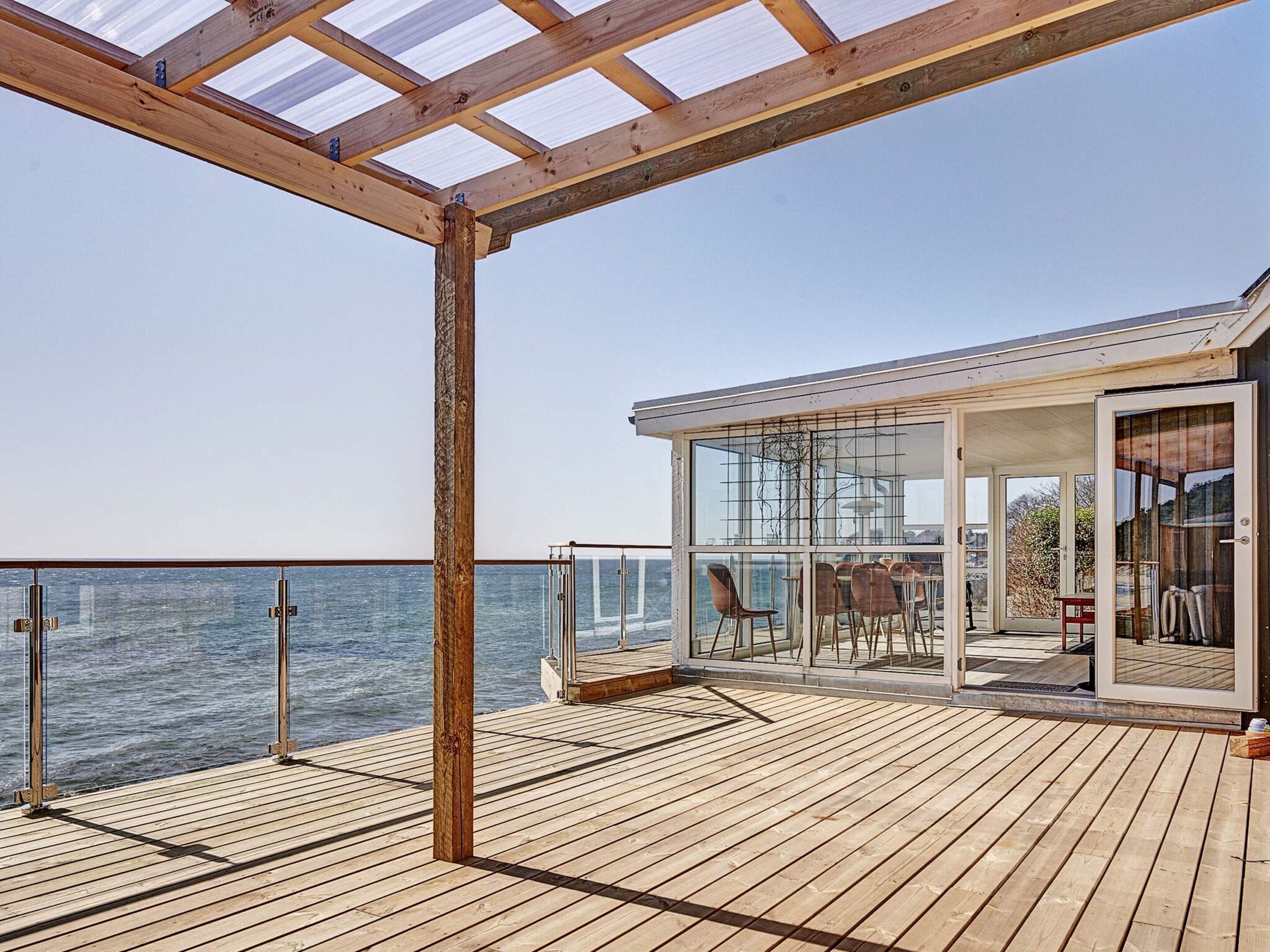 Une terrasse en bois avec un espace repas fermé par des baies vitrées, offrant une vue sur la mer.
