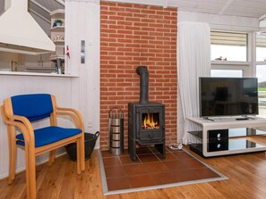 A living room with a brick fireplace, a blue chair, a TV on a stand, and a window with a view. - 8 Person Holiday Home in Nordborg-by Traum (Nordborg)