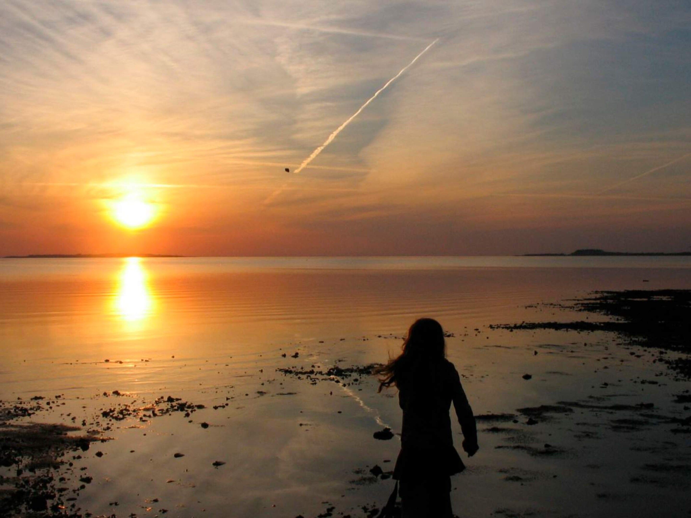 A silhouette of a person walking on a beach during sunset with a clear sky and a visible contrail.
